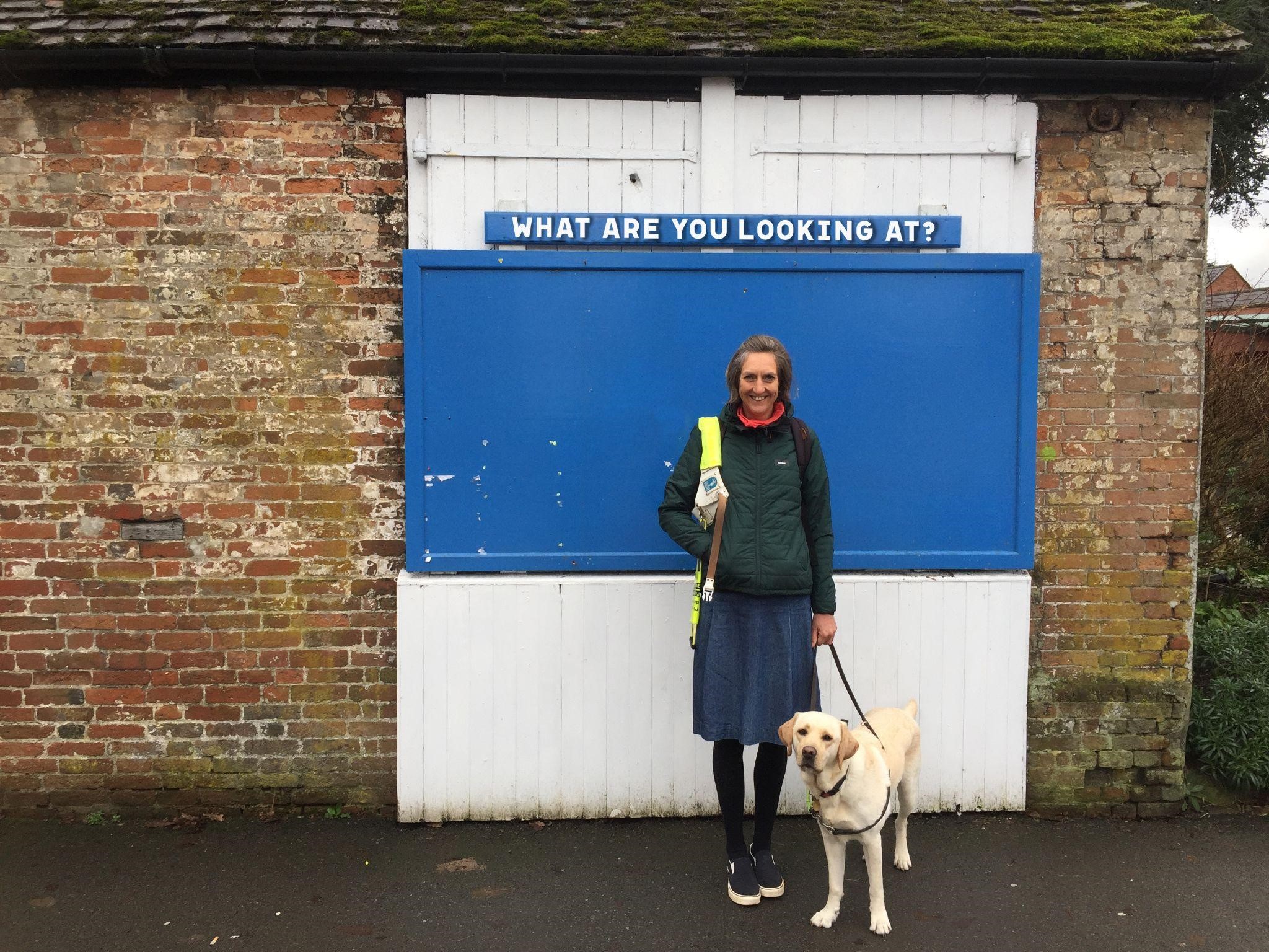 Angela Charles with guide dog (a golden Labrador) in front of a brick building with a blue & white door